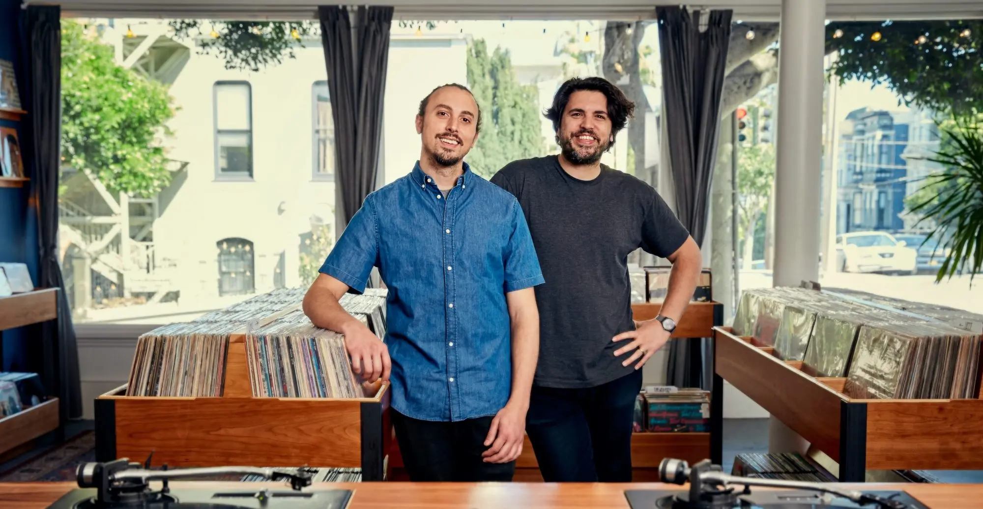 Two owners standing inside a bright Mission District record store with sunlight, large front windows, and rows of neatly organized vinyl bins.