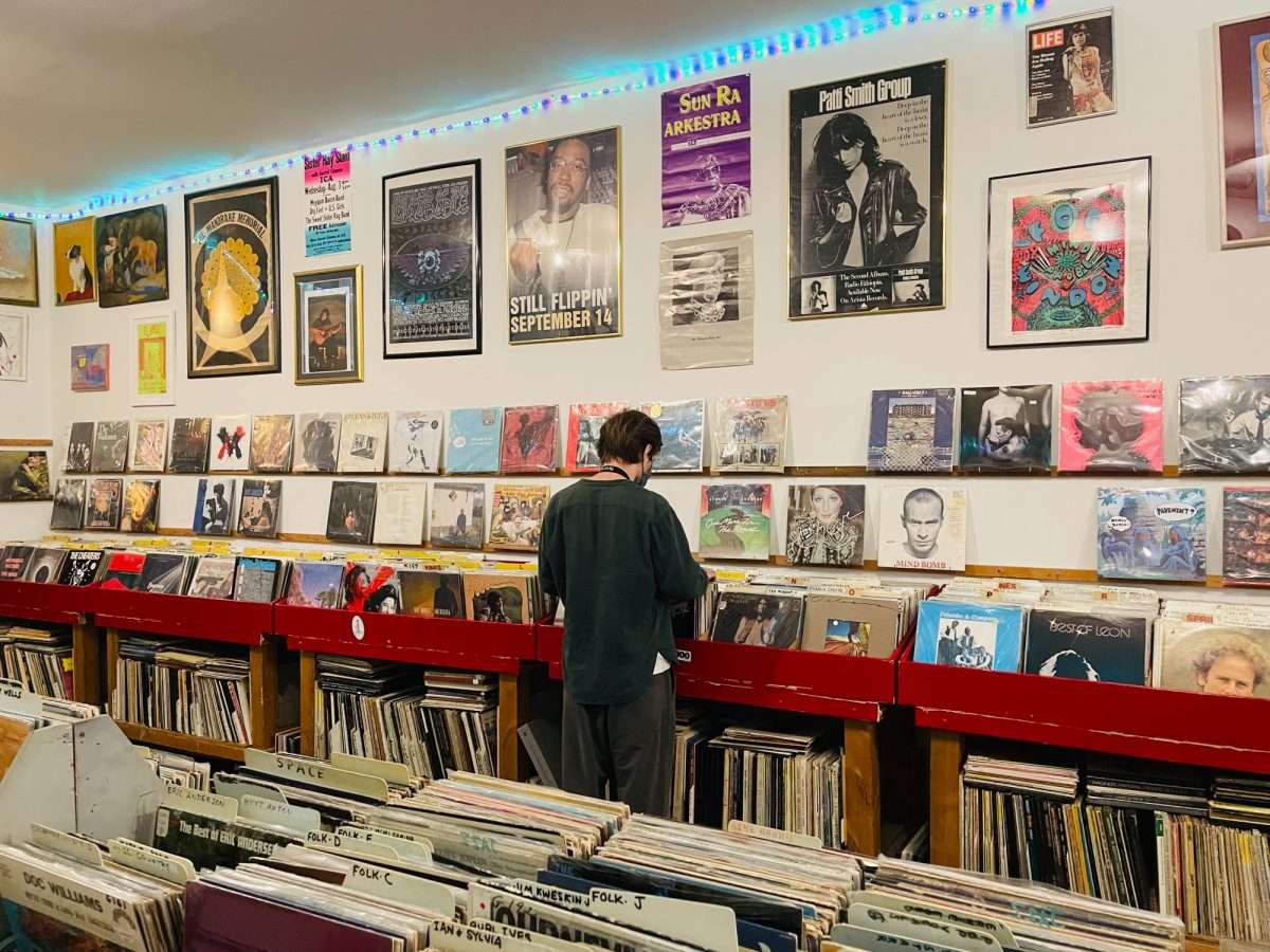 A person browsing through vinyl records inside Repo Records in Philadelphia, surrounded by red record bins and walls covered in framed posters of artists like Patti Smith and Sun Ra under colorful LED lights.