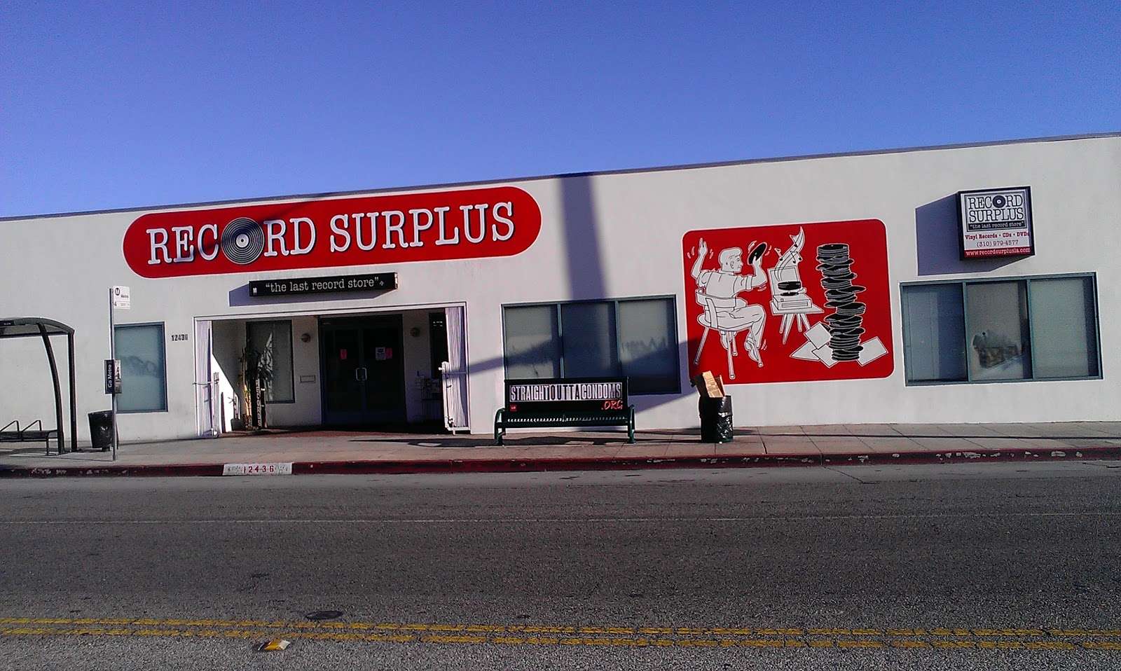 Exterior of Record Surplus in Los Angeles, featuring its signature red-and-white mural of a cartoon character surrounded by vinyl stacks. The shop’s tagline, “The Last Record Store,” is displayed above the entrance under a clear blue sky.
