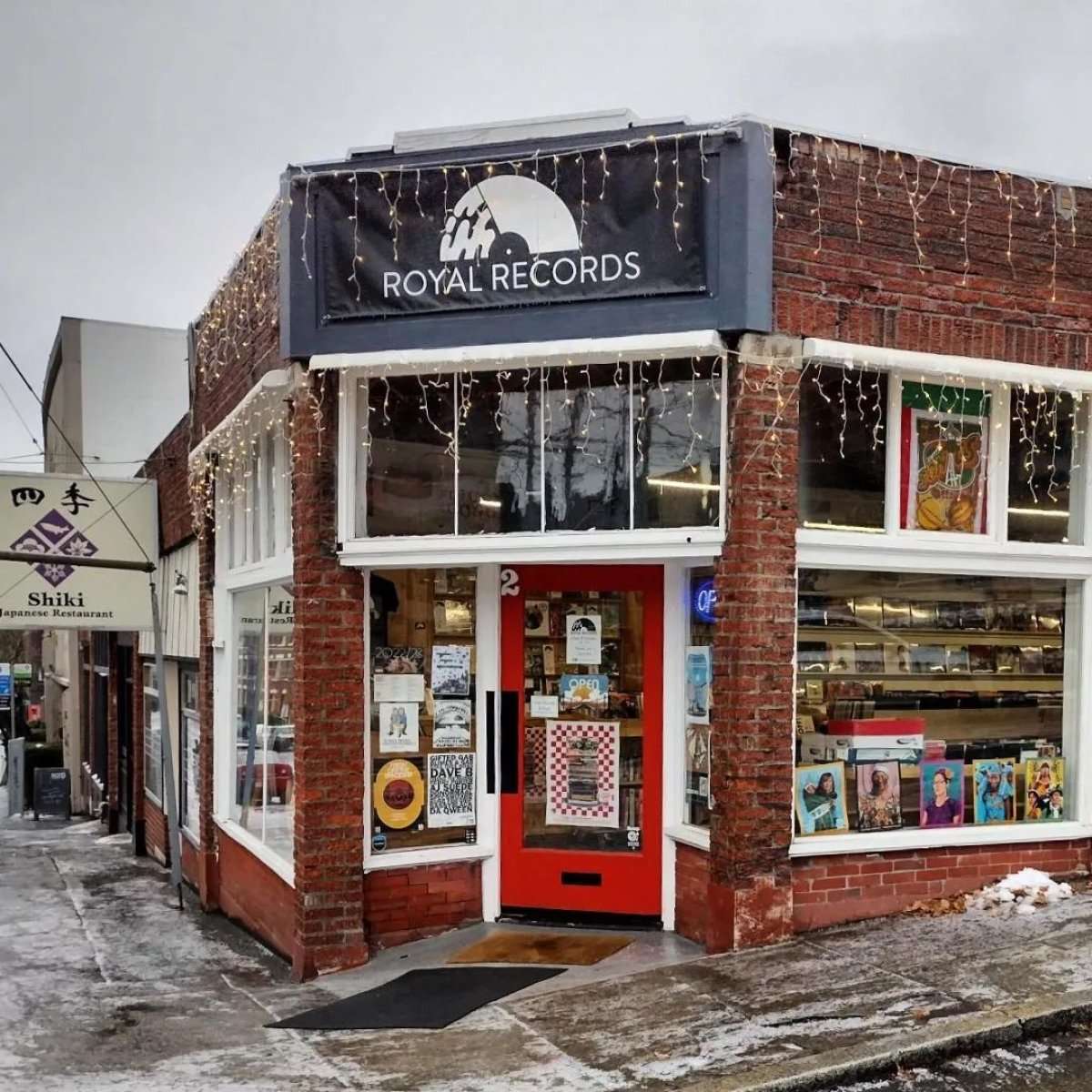 Brick corner shop with a red door, string lights, and window displays of vinyl at Royal Records on a winter day.