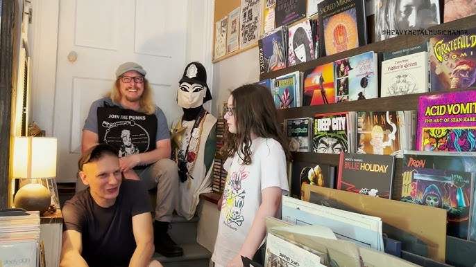 Inside Sisters in Christ Records, an intimate New Orleans record store with walls of punk, jazz, and experimental vinyls, and three people chatting on the steps surrounded by album covers.