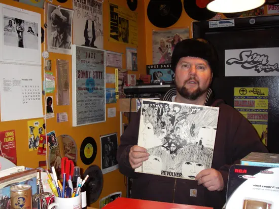 Clerk holding a Beatles “Revolver” LP at a cozy, poster-covered counter inside the shop.