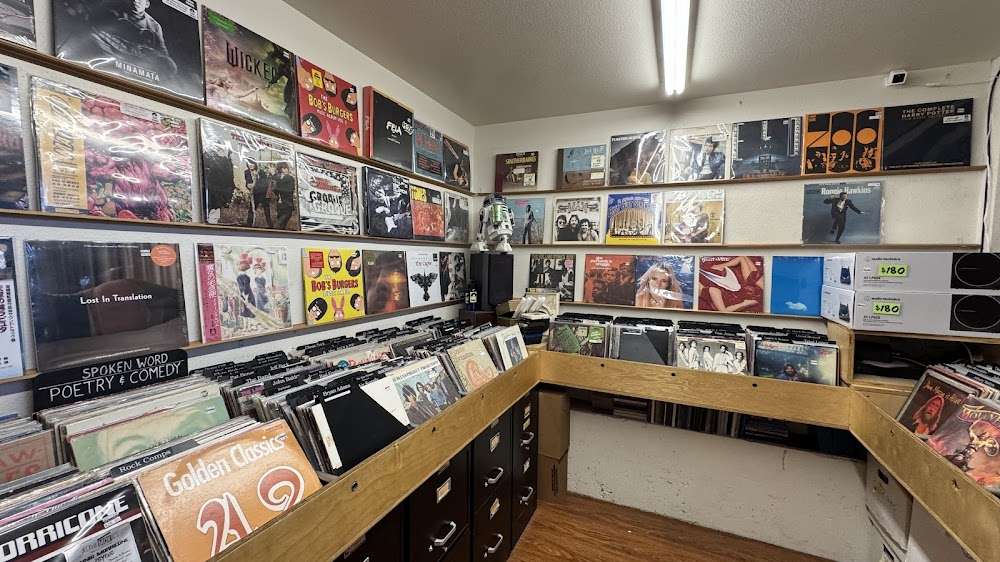 L-shaped wooden bins packed with LPs, wall ledges displaying featured album jackets, and bright fluorescent lighting in a cozy corner room.