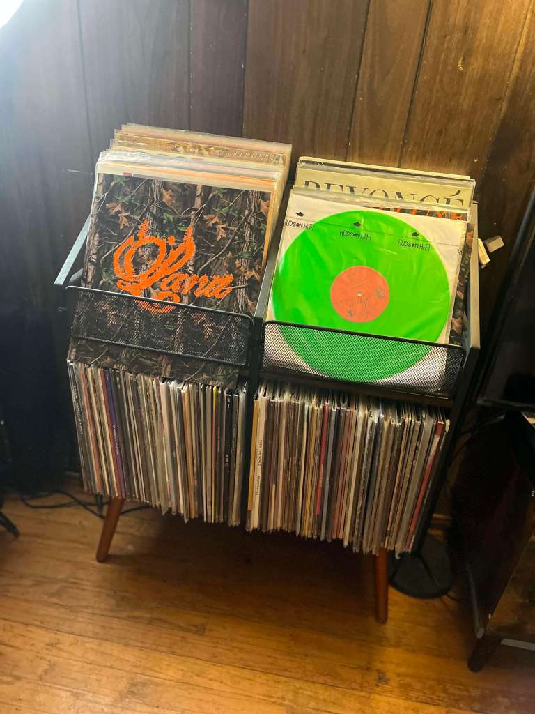 A mid-century style wooden record stand with two black mesh bins filled with vinyl records. One visible record has a bright neon green disc, while others include colorful album art. The stand sits against a wood-paneled wall on hardwood flooring, showcasing an organized and stylish vinyl collection.