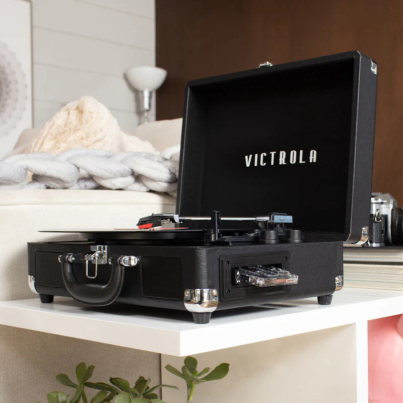 Black Victrola suitcase-style record player with lid open and a vinyl spinning, sitting on a white shelf in a cozy living room; chrome corners and handle visible with a plant and sofa in the background.