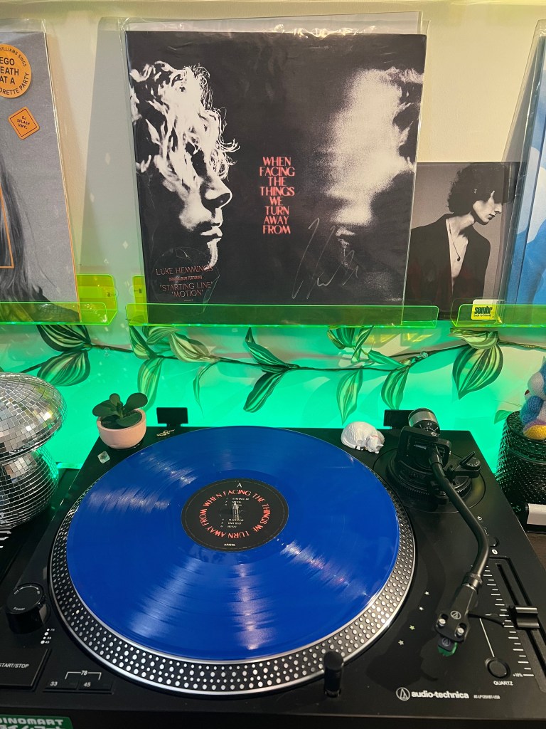 Close-up of a blue vinyl record spinning on an Audio-Technica turntable, with Luke Hemmings’ When Facing the Things We Turn Away From album cover displayed on a neon-green acrylic shelf against a leafy, green-lit backdrop.