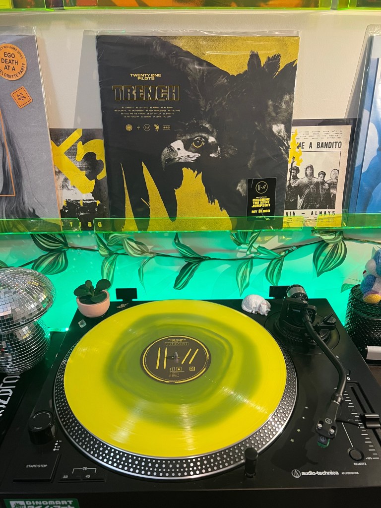Yellow translucent copy of Twenty One Pilots’ Trench spinning on an Audio-Technica turntable, with the black-and-yellow album cover displayed above on a neon green acrylic shelf, surrounded by green leaf lighting, a disco ball, and small desk decor.