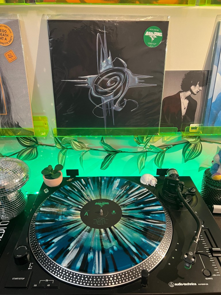 Close-up of a teal-and-black splatter vinyl spinning on an Audio-Technica turntable, with Bad Suns’ “Accelerator” cover on a neon green wall shelf and green LED backlighting with leafy garland.