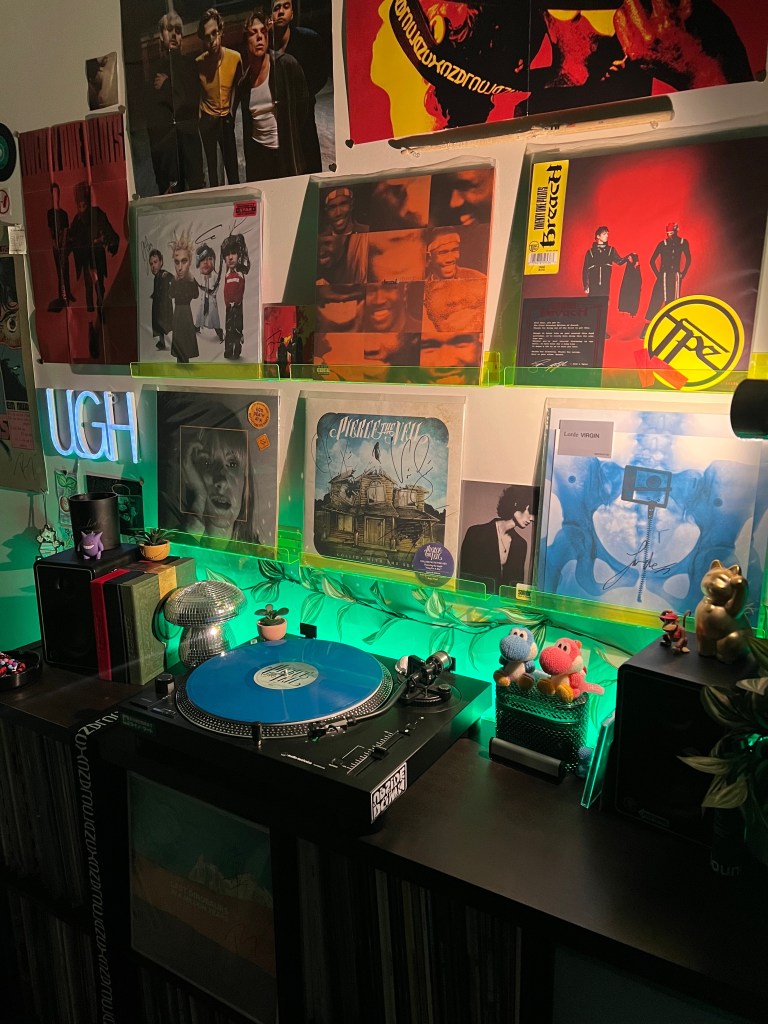 Cozy, dimly lit vinyl setup with green LED backlighting: a black DJ-style turntable spinning a bright blue record sits on a dark shelving unit, surrounded by small plants, a disco-mushroom ornament, Yoshi plush figures, a Maneki-neko statue, and compact speakers, with neon “UGH” sign, colorful album sleeves, and posters filling the wall above.