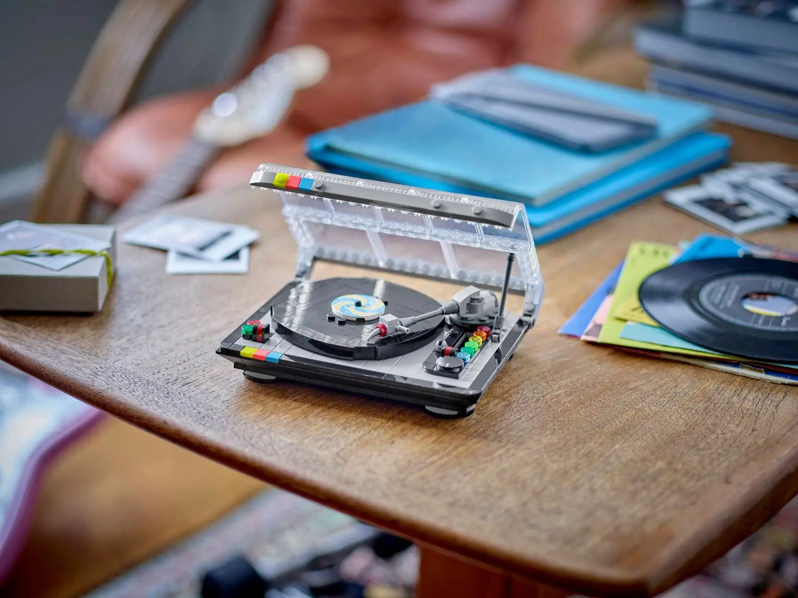 The LEGO Retro Record Player (40699) sits on a wooden side table with its transparent dust cover propped open, revealing a black vinyl record with a printed label and a detailed brick-built tonearm. Colorful button details accent the body. Real vinyl records, album sleeves, and Polaroid photos are scattered around it in the background.