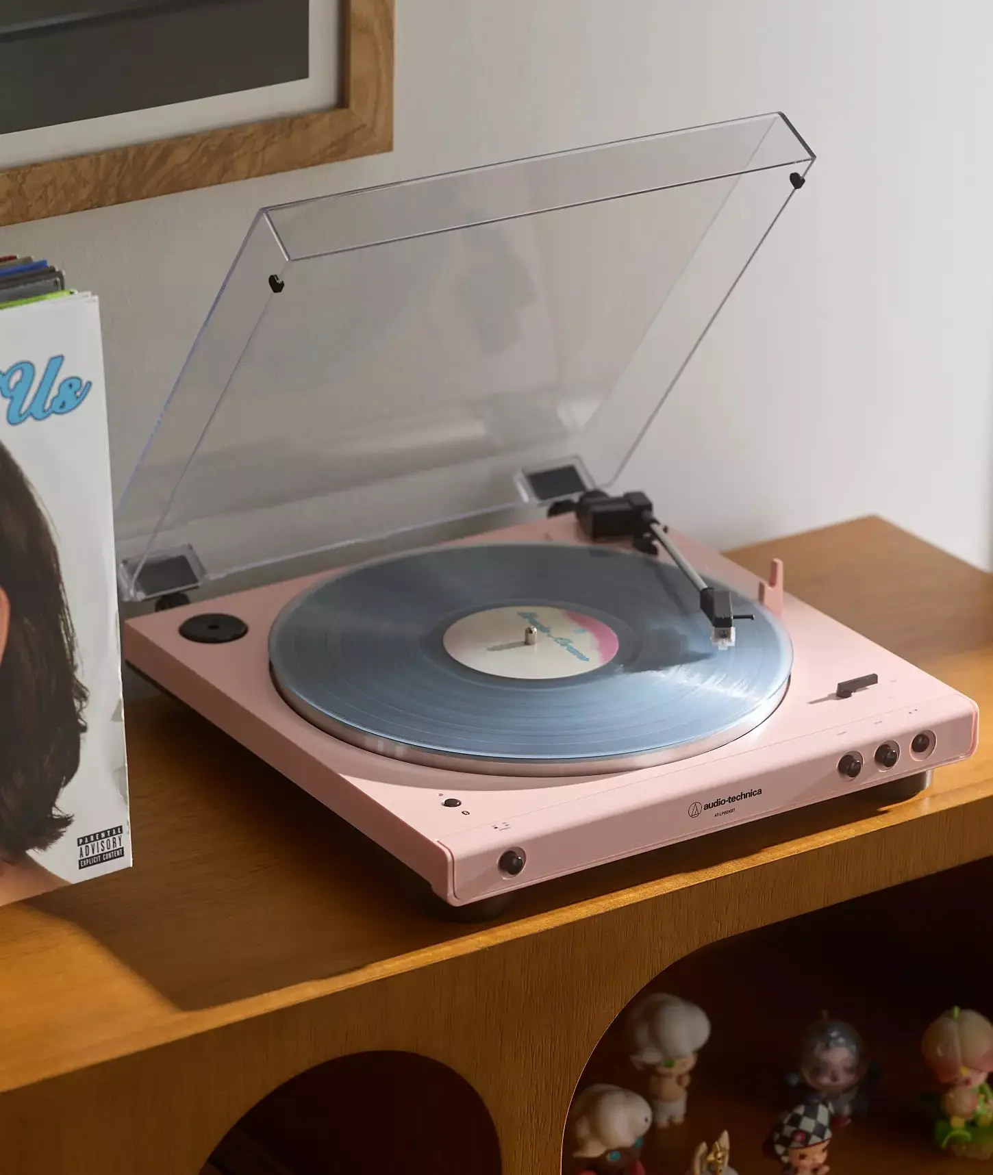 Pink Audio-Technica AT-LP60XBT turntable with dust cover open, playing a blue marbled vinyl record, on a wooden shelf beside a record sleeve and collectible figurines.