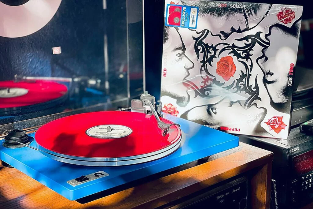 A blue U-Turn Orbit Plus turntable sits on a wooden cabinet in warm sunlight, playing a translucent red vinyl record. Behind it stands the shrink-wrapped Blood Sugar Sex Magik cover, with black tribal-style artwork, red roses, and a Walmart-exclusive red vinyl sticker. A Sony stereo receiver is visible at right, and the clear dust cover in the background reflects a second red record.